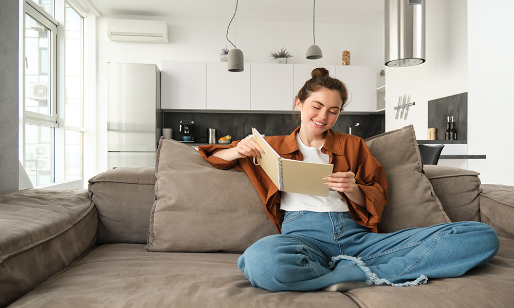 Smiling young brunette woman, sits on sofa in living room, holds notebook, reads her notes, studies for exam, student does her homework at home.